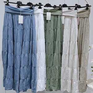 May include: Five tiered maxi skirts in various colours hang on a clothing rack. The skirts are in shades of blue, white, olive green, off-white, and tan. Each skirt has a wide, folded waistband and a flowing, tiered design.