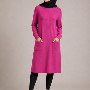 May include: A long-sleeved magenta tunic dress featuring a round neckline, two front patch pockets, and a subtle diamond-patterned texture on the upper chest. The garment is paired with black leggings and black open-toed slide sandals.
