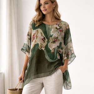 May include: A sheer, green floral top with an asymmetrical hemline. The top features a pattern of large, cream-coloured flowers with red and green accents. The sleeves are short and wide. The model is wearing white trousers.