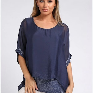 Puede incluir: Una blusa azul marino con cuello redondo y mangas cortas. La blusa tiene un diseño en capas, con una capa exterior transparente y un ribete de lentejuelas en las mangas y el dobladillo. La modelo lleva pantalones de color claro.