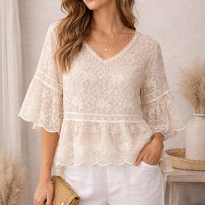 Puede incluir: Top de encaje color marfil con escote en V y mangas acampanadas. El top presenta detalles florales y un dobladillo festoneado. La modelo lleva pantalones blancos y sostiene un pequeño bolso de mano tejido. El estilo es femenino y elegante.