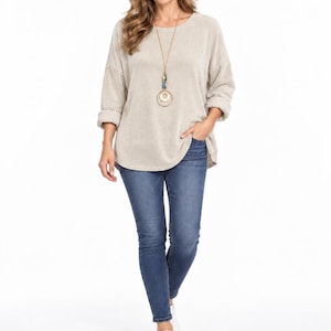 Peut inclure: Un haut beige à manches longues avec une encolure ronde et des poignets retroussés, associé à un jean bleu et des baskets blanches. Un long collier doré présente un pendentif avec des perles colorées et deux anneaux circulaires suspendus.