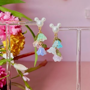 May include: A pair of whimsical earrings featuring a white pearl-like stud with a gold-coloured post. Each earring has a cluster of pastel-coloured flower charms in shades of blue, green, and white, with small green leaf accents. The earrings are displayed against a pink background.