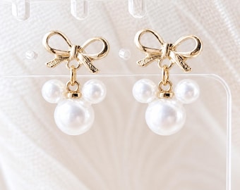 May include: A pair of gold-toned earrings featuring a bow at the top and a dangling charm composed of three white spherical faux pearls arranged in a circular pattern, resembling a mouse head silhouette with two smaller pearls as ears.
