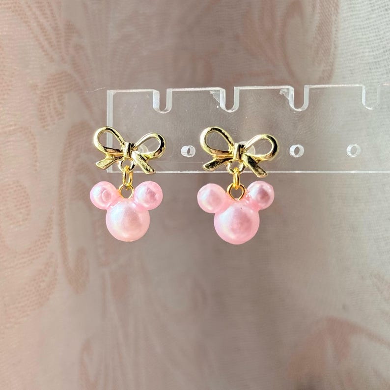 K&ouml;nnte beinhalten: Ein Paar goldfarbene Schleifenohrringe mit rosa, perlenartigen Mickey Mouse-Kopf-Anh&auml;ngern. Die Ohrringe h&auml;ngen an einem transparenten Pr&auml;sentationsst&auml;nder vor einem weichen rosa Hintergrund. Ein niedliches und skurriles Accessoire.