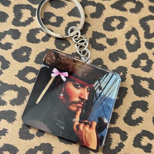 Pu&ograve; includere: Portachiavi quadrato con un ritratto di un personaggio con cappello e bandana rossa, con un fiocco rosa. Il portachiavi ha un anello e una catena in metallo argentato. Lo sfondo &egrave; un motivo leopardato.
