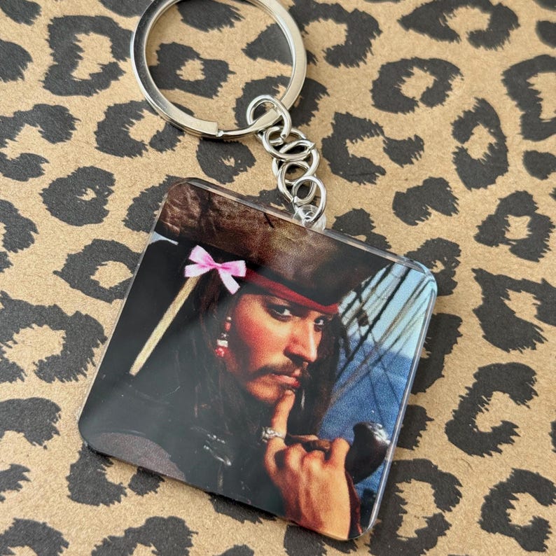 Pu&ograve; includere: Portachiavi quadrato con un ritratto di un personaggio con cappello e bandana rossa, con un fiocco rosa. Il portachiavi ha un anello e una catena in metallo argentato. Lo sfondo &egrave; un motivo leopardato.