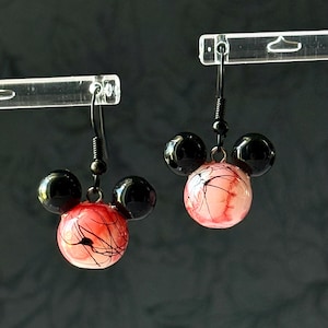 Puede incluir: Un par de pendientes colgantes de diseño caprichoso. Cada pendiente presenta una esfera central roja y blanca con líneas negras, flanqueada por dos esferas negras, creando una silueta de ratón. Los pendientes cuelgan de ganchos negros.