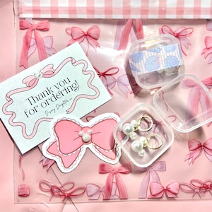 K&ouml;nnte beinhalten: Eine rosa, transparente Plastikt&uuml;te mit einem rosa Schleifenmuster, die eine Karte mit der Aufschrift "Thank you for ordering!" und einen rosa Schleifenaufkleber enth&auml;lt. Au&szlig;erdem ein kleiner, klarer Beh&auml;lter mit Ohrringen und ein kleiner Beh&auml;lter mit Muschelschalen-Aufklebern.