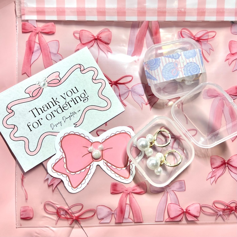 K&ouml;nnte beinhalten: Eine rosa, transparente Plastikt&uuml;te mit einem rosa Schleifenmuster, die eine Karte mit der Aufschrift "Thank you for ordering!" und einen rosa Schleifenaufkleber enth&auml;lt. Au&szlig;erdem ein kleiner, klarer Beh&auml;lter mit Ohrringen und ein kleiner Beh&auml;lter mit Muschelschalen-Aufklebern.