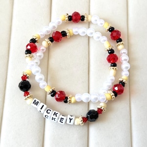 May include: Two beaded bracelets with white, red, black, and yellow beads. One bracelet has the word "MICKEY" spelled out in white letter beads. The bracelets have gold-colored accents and are on a white surface.