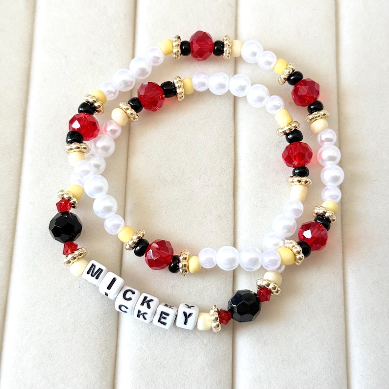 May include: Two beaded bracelets with white, red, black, and yellow beads. One bracelet has the word "MICKEY" spelled out in white letter beads. The bracelets have gold-colored accents and are on a white surface.