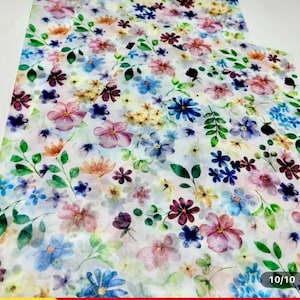 May include: Floral patterned fabric featuring a watercolor design of various flowers in shades of pink, blue, purple, and yellow. Green leaves and stems are interspersed throughout the design, creating a vibrant and colorful pattern. The fabric has a white background.