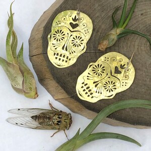 May include: A pair of gold-tone metal earrings shaped like sugar skulls with intricate cut-out designs. The earrings have a hammered texture and feature small hearts and floral details.