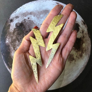 May include: A pair of gold lightning bolt earrings with a hammered texture. The earrings are dangling and have a silver hook closure.