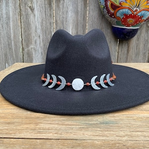 May include: A black felt fedora hat with a brown leather band adorned with silver crescent moon charms and red beads.