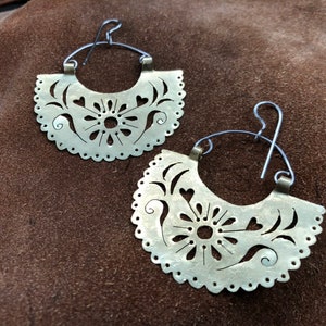 May include: A pair of silver hoop earrings with a delicate, intricate floral design. The earrings are made of metal and have a cutout pattern.