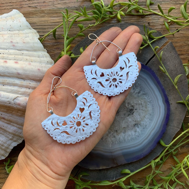 May include: A pair of white paper earrings with a delicate floral design and cut-out hearts. The earrings are silver-toned and have a hoop style.