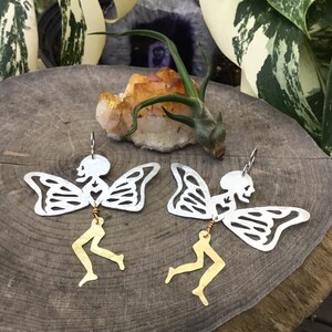 Pode incluir: Um par de brincos de prata com um design de fada esqueleto com pernas douradas. As fadas t&ecirc;m asas de borboleta e um cora&ccedil;&atilde;o no peito.