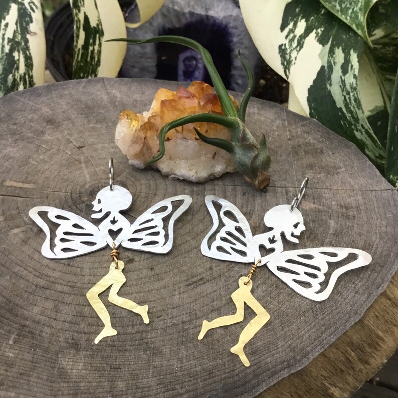 Pode incluir: Um par de brincos de prata com um design de fada esqueleto com pernas douradas. As fadas t&ecirc;m asas de borboleta e um cora&ccedil;&atilde;o no peito.