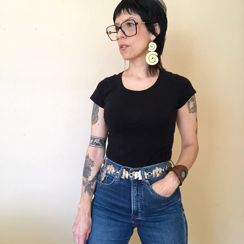 May include: A person wearing a black t-shirt with a silver chain belt and a brown leather bracelet. They have a tattoo on their arm and are wearing blue jeans.