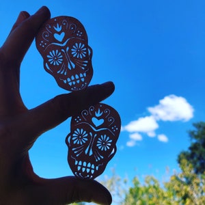 May include: Two paper cutouts of sugar skulls, a traditional Mexican symbol, against a blue sky background. The skulls are white and feature intricate designs, including hearts, flowers, and swirls.