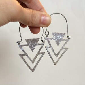 May include: Silver triangle earrings with a hammered texture. The earrings are made of two triangles, one inside the other, and are suspended from a wire hoop.