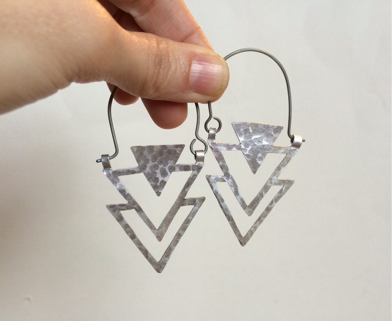 May include: Silver triangle earrings with a hammered texture. The earrings are made of two triangles, one inside the other, and are suspended from a wire hoop.