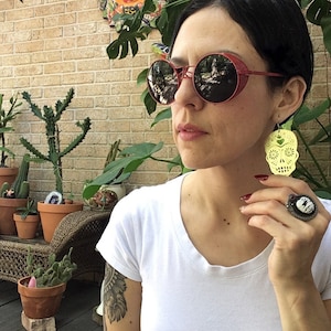 May include: A woman wearing round red sunglasses with a yellow sugar skull earring. The earring is a cutout design with a heart in the center of the skull.