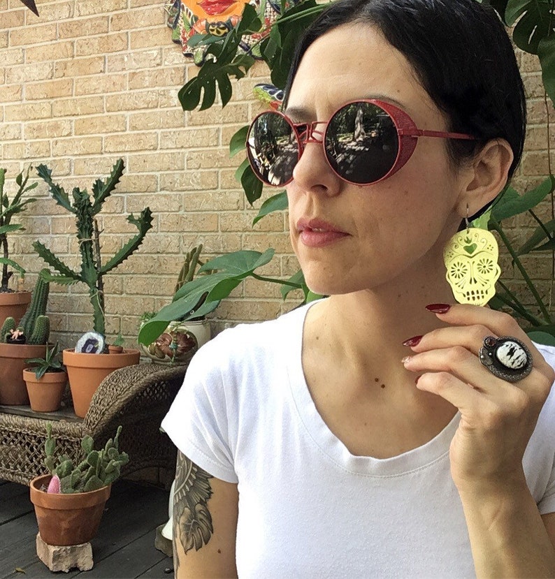 May include: A woman wearing round red sunglasses with a yellow sugar skull earring. The earring is a cutout design with a heart in the center of the skull.