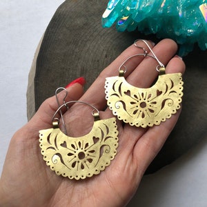 May include: A pair of gold-toned earrings with a delicate, intricate design. The earrings are shaped like a half-circle and feature a floral pattern with cut-out details.
