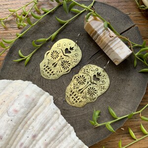 May include: A pair of brass earrings shaped like sugar skulls with intricate cutouts and a hammered texture. The earrings are on a wooden surface with a white and brown crystal.