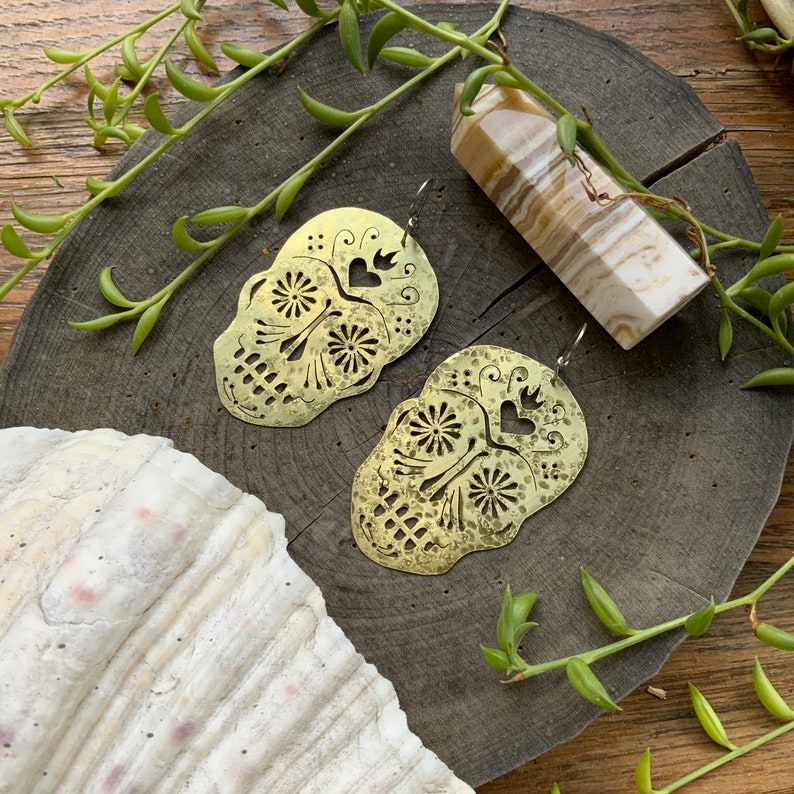 May include: A pair of brass earrings shaped like sugar skulls with intricate cutouts and a hammered texture. The earrings are on a wooden surface with a white and brown crystal.