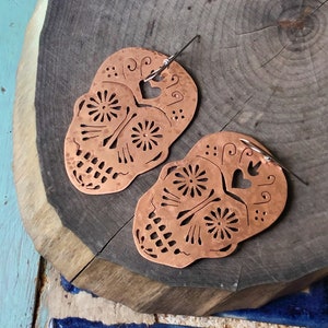 May include: A pair of copper earrings shaped like sugar skulls with intricate cutouts and a hammered texture.