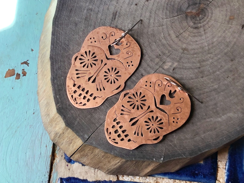 May include: A pair of copper earrings shaped like sugar skulls with intricate cutouts and a hammered texture.