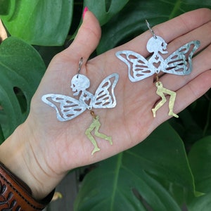 Pode incluir: Um par de brincos de borboleta esqueleto prateados com pernas douradas penduradas. Os brincos s&atilde;o feitos &agrave; m&atilde;o e t&ecirc;m um design delicado e caprichoso.