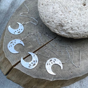 May include: Four silver crescent moon earrings with delicate floral cutouts and dangling chains. The earrings are displayed on a wooden surface.