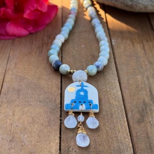 May include: A handcrafted necklace featuring a pendant with a blue and white design, accented with gold wire and clear teardrop-shaped stones. The necklace has a beaded strand of light blue and gray stones.