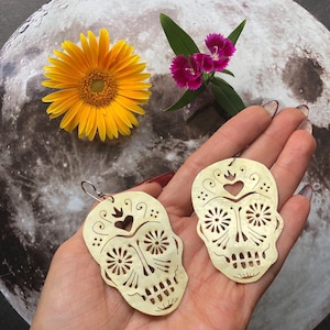 May include: A pair of gold-colored earrings shaped like sugar skulls with intricate cut-out designs. The earrings have a heart and flower details.