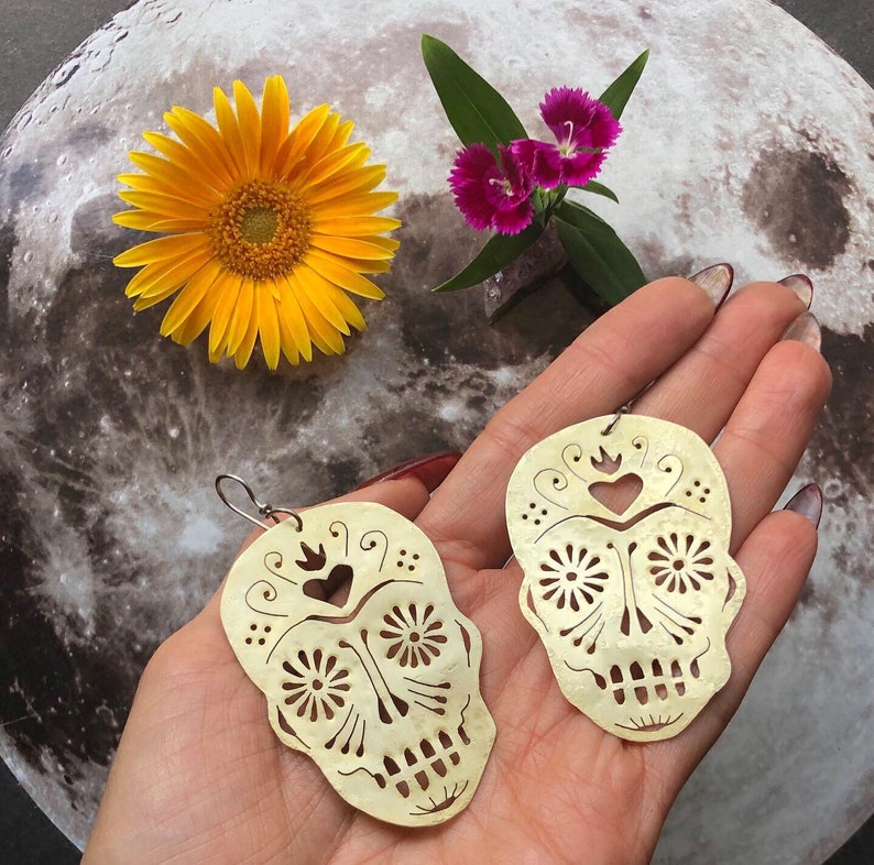 May include: A pair of gold-colored earrings shaped like sugar skulls with intricate cut-out designs. The earrings have a heart and flower details.