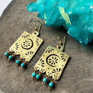 May include: Gold earrings with a cutout design featuring a crescent moon and a flower. The earrings have turquoise beads dangling from the bottom.