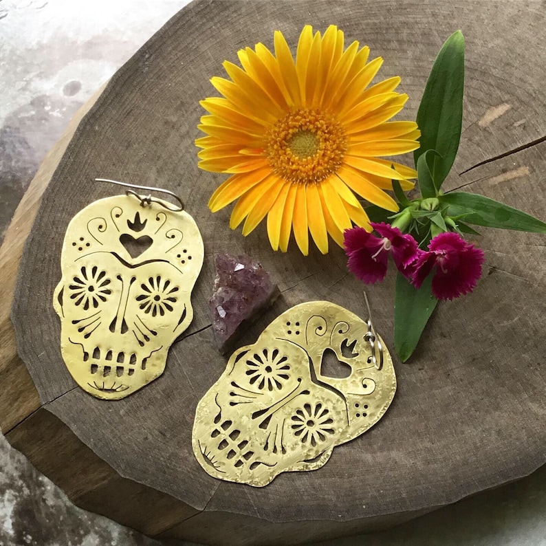 May include: A pair of brass earrings shaped like sugar skulls with intricate cutouts. The earrings are on a wooden surface with a yellow flower and a purple crystal.