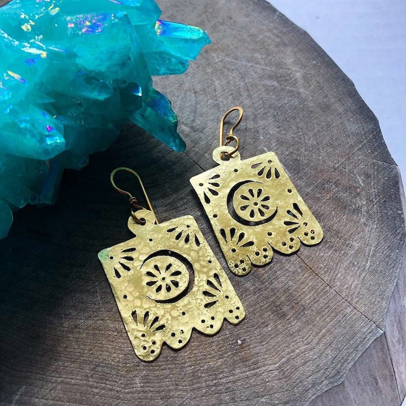 May include: A pair of gold-toned earrings with a crescent moon and floral design. The earrings are made of brass and have a hammered texture.