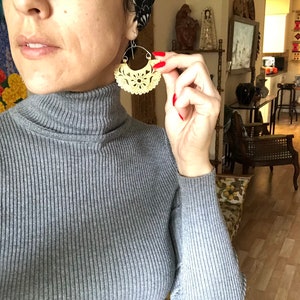 May include: A woman wearing a gray turtleneck sweater with a large, intricate, cream-colored, carved wooden earring. The earring is shaped like a crescent moon with a floral design.