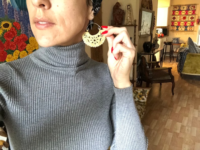 May include: A woman wearing a gray turtleneck sweater with a large, intricate, cream-colored, carved wooden earring. The earring is shaped like a crescent moon with a floral design.