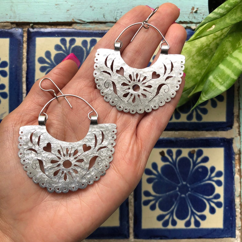 May include: A pair of silver hoop earrings with a cut-out design featuring a flower and hearts. The earrings are handmade and have a hammered texture.