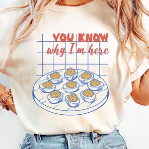 Puede incluir: Camiseta color marfil con el texto "YOU KNOW why I'm here" en rojo y azul. Debajo, un gráfico de huevos rellenos en un plato. Una prenda casual para el uso diario.
