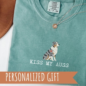 Peut inclure: T-shirt vert clair avec un dessin d'un berger australien et le texte "KISS MY AUSS". Un collier doré avec un pendentif est posé sur le t-shirt. Les mots "PERSONALIZED GIFT" sont sur une bannière en dessous.