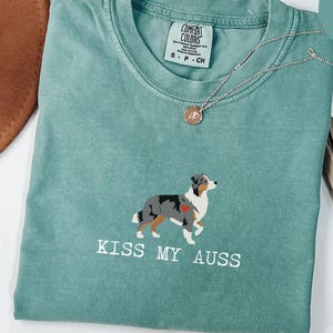 May include: Light green Comfort Colors t-shirt with a dog graphic and the text "KISS MY AUSS". The dog is a mix of gray, white, and brown with a red heart. A gold necklace with a pendant is on the shirt.