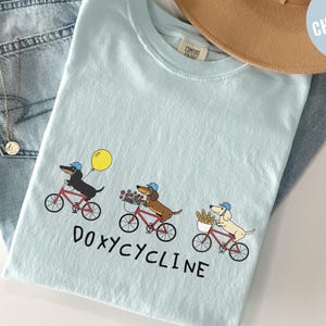 May include: Light blue t-shirt featuring a whimsical illustration of three dachshunds riding bicycles. Each dog wears a blue cap and carries items like a balloon, flowers, and bread. The word "DOXYCYCLINE" is printed below the dogs.